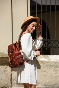 brunette with long hair wearing white dress and beige hat posing with luxury leather backpack. old city street. Vacation, tourism and travel