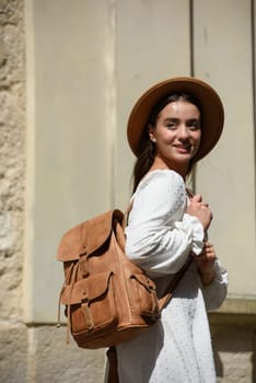 brunette with long hair wearing white dress and beige hat posing with luxury leather backpack. old city street. Vacation, tourism and travel