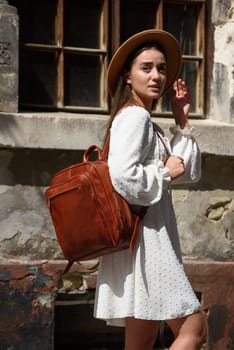 brunette with long hair wearing white dress and beige hat posing with luxury leather backpack. old city street. Vacation, tourism and travel