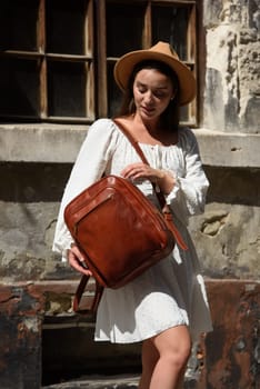 brunette with long hair wearing white dress and beige hat posing with luxury leather backpack. old city street. Vacation, tourism and travel