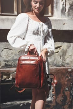 brunette with long hair wearing white dress and beige hat posing with luxury leather backpack. old city street. Vacation, tourism and travel