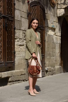 pretty brunette girl in satin long dress ankle strap shoes and checkered jacket posing with leather handbag outside.