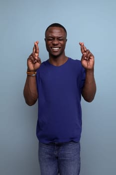 cute young african man in blue t-shirt crossed fingers on blue background.