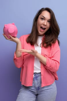 positive brunette young woman in a shirt and jeans holding a piggy bank with savings.