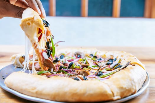 Hand taking a slice of supreme pizza on wooden table. Close-up of a hand taking a delicious slice of supreme pizza with vegetables