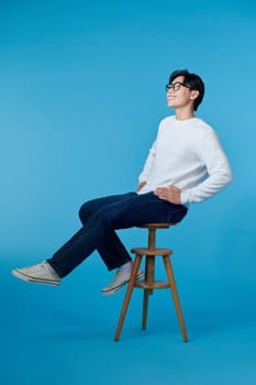Feeling comfortable. Joyful positive young man sitting on a stool and smiling isolated on blue.