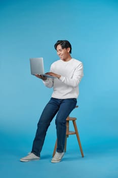 Handsome emotional man sitting on stool and using laptop isolated on blue background