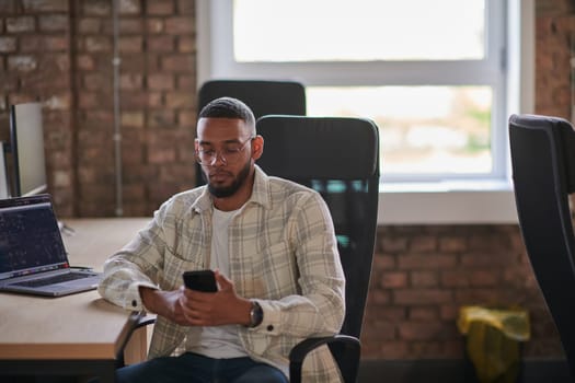 A young African American entrepreneur takes a break in a modern office, using a smartphone to browse social media, capturing a moment of digital connectivity and relaxation amidst his business endeavors
