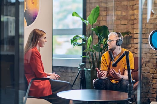 A gathering of young business professionals, some seated in a glass-walled office, engage in a lively conversation and record an online podcast, embodying modern collaboration and dynamic interaction.