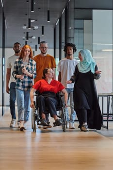 A diverse group of young business people walking a corridor in the glass-enclosed office of a modern startup, including a person in a wheelchair and a woman wearing a hijab, showing a dynamic mix of innovation and unity