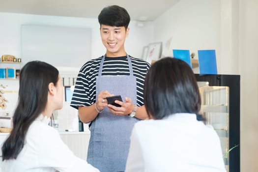 Cheerful smiling waiter in apron using table while take order and talk to clients cafe coffee shop visitors, friendly professional man server wear apron write menu choice, serving staff good customer service.