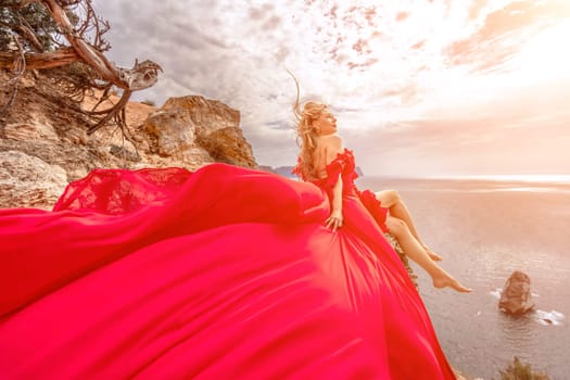 woman red silk dress sits by the ocean with mountains in the background, her dress swaying in the wind
