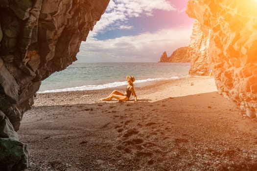 Woman travel sea. View of a woman in a black swimsuit from a sea cave Attractive woman enjoying the sea air sits on the beach and looks at the sea. Behind her are rocks and the sea.