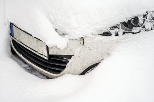 Car under deep layer of snow after heavy snowstorm, detail on only part of headlight and front bumper visible.