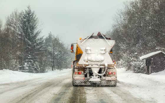 Snow covered orange highway maintenance gritter truck on slippery road, heavy snowing and trees in background.