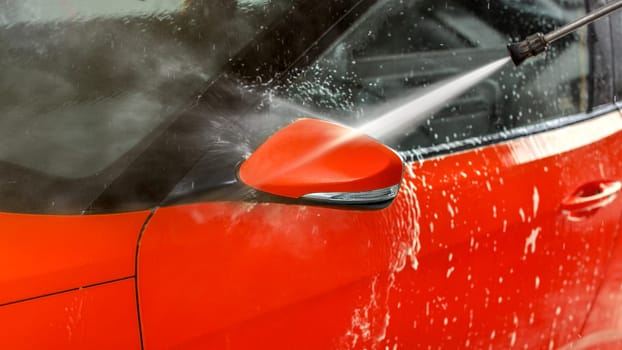 Side mirror of red car washed in self service carwash, jet water spraying from nozzle