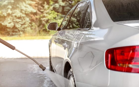 Silver car washed in self serve carwash, jet water spraying from nozzle to wheels, sun lit trees in background.