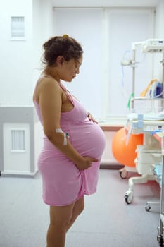 Side portrait of a pregnant woman, birthing mother holding her belly while having contraction during labor process, in hospital ward. Pregnancy. Easy delivery. Childbirth. Obstetrics and gynecology
