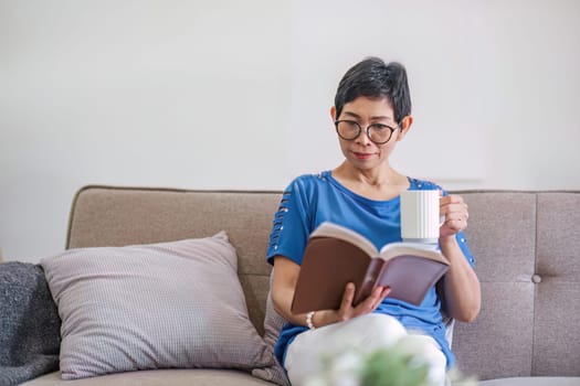 A happy retired 60s Asian woman reading a book while having her coffee on a sofa at home..