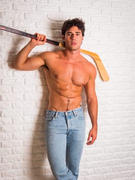 Handsome young man shirtless with hockey stick in hands leaning against white brick wall, looking at camera, wearing jeans