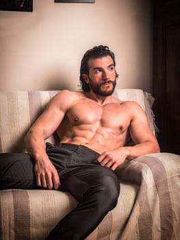 Handsome muscular young man at home laying on couch with shirt open on naked torso, an expression of tranquility, looking away, in a health and fitness concept