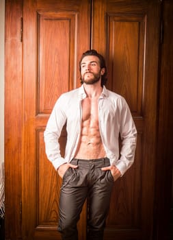 Sexy handsome man standing in white open shirt with a smile in front of wood closet doors