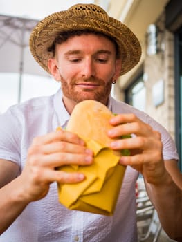 A man in a straw hat is holding a sandwich outdoor, sitting and looking at the food.