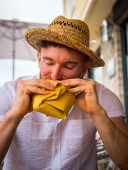 A man in a straw hat is holding a sandwich outdoor, sitting and looking at the food.