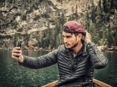 A man sitting in a boat holding a cell phone. Photo of a man enjoying the scenic beauty of the Dolomites in Italy while using his cell phone in a boat