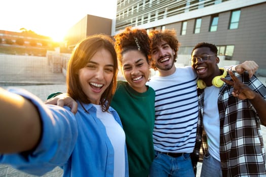Group of happy and smiling multiracial friends taking selfie and having fun together outdoors in the city during sunset. Looking at camera. Friendship and technology concept.