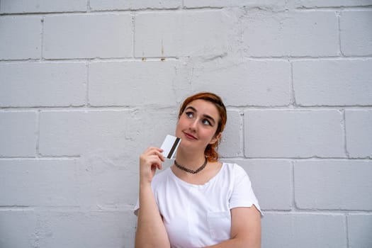 Positive young Hispanic female in makeup holding credit card with magnetic strip in fingers near chin and looking up while standing with crossed hand near brick wall in daylight