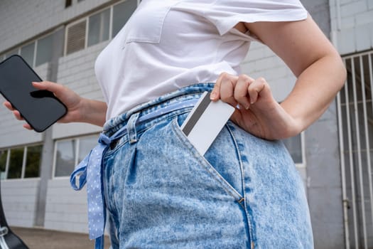 From above of anonymous young female in jeans and white shirt standing with smartphone outside building with windows in daylight while putting card with magnetic strip in pants pocket.