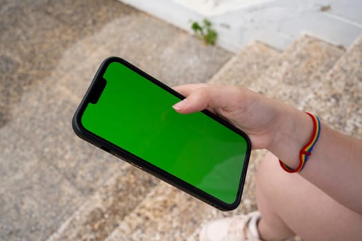 From above of anonymous young female in sneakers with rainbow hand band sitting on marble steps with chromakey screen mobile phone and checking information while browsing messages