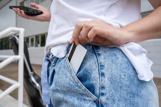 From above of anonymous young female standing with smartphone near guitar case outside building with windows in daylight while putting credit card with magnetic strip in jean pants pocket