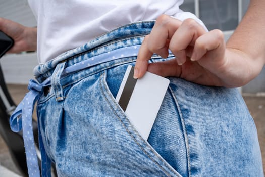 Anonymous young female putting credit card with magnetic strip in jean pants pocket with manicured fingers and holding mobile phone while standing near guitar case in daylight