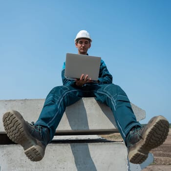 Caucasian male builder in hardhat sits on floor slabs and uses laptop at construction site