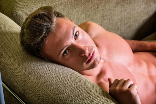 A shirtless man laying on a couch. Photo of a handsome blond man with muscular physique lounging on a couch looking at camera