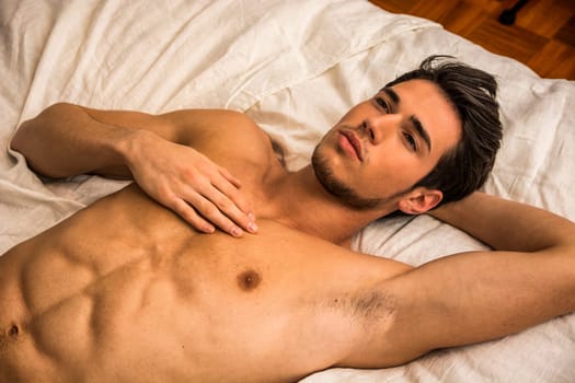 A shirtless man laying on a bed with white sheets. Photo of a shirtless man relaxing on a bed with white sheets looking up