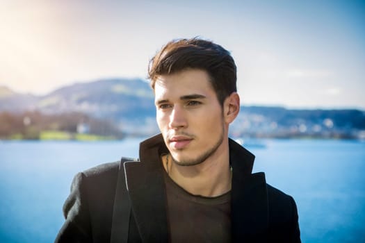 A young man standing in front of a body of water, looking to a side