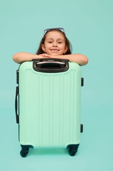 Adorable Caucasian little child girl, traveler, tourist, passenger, going for weekend getaway, smiling looking at camera, posing with blue suitcase, isolated over studio background. Copy ad space