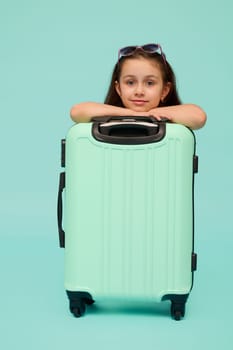 Beautiful Caucasian little child girl, traveler, tourist, passenger, going for weekend getaway, looking confidently at camera, posing with blue suitcase, isolated over studio background. Copy ad space