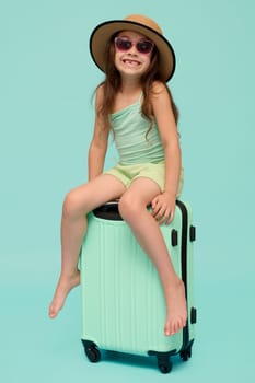 Full length vertical studio portrait of a Caucasian cute child girl 6 years old, traveler, passenger in sunglasses and straw summer hat, looking at camera, travelling abroad, sitting on her suitcase