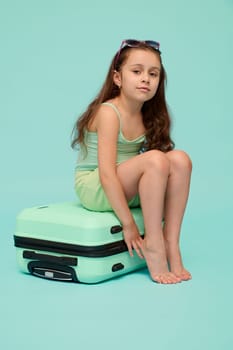 Full length portrait of a little traveler girl dressed in blue turquoise trendy in summer wear, sitting on a plastic suitcase, smiling cutely looking at camera, isolated studio background