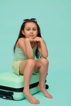 Adorable little kid girl, traveler tourist, passenger, dressed in blue turquoise trendy in summer wear, sitting on a plastic suitcase, smiling cutely looking at camera, isolated studio background