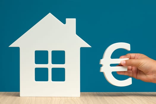 The value of real estate in Europe. Buying or selling a home for Euro. Model of a white house and a hand with a euro symbol on a blue background close-up. copy space. High quality photo