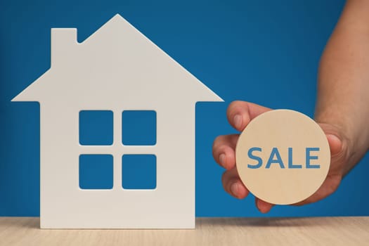 House sale. Model of the White House as a symbol of the sale of real estate. Close-up hand holding a wooden sign with the inscription SALE. Banner on the theme of real estate on a blue background