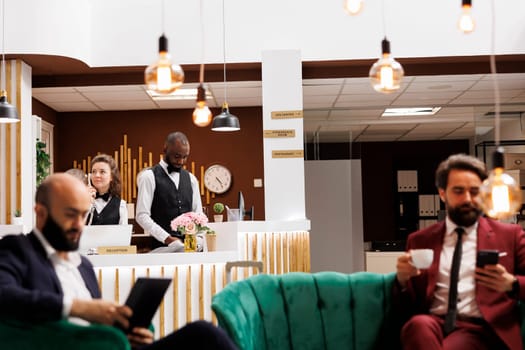 Diverse hotel staff works at front desk to assist guests and provide excellent concierge services in reception lobby. Receptionist and bellboy in lobby, greeting travellers on journey.