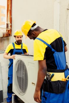 Precise serviceman and african american colleague installing new external hvac system for client. Meticulous wiremen optimizing new air conditioner performance, ensuring it operates at productivity