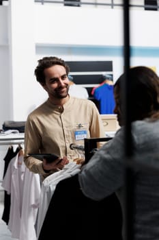 Shopping mall smiling man employee assisting client in choosing fashionable outfit. Clothing store consultant holding digital tablet and offering customer help in selecting casual apparel