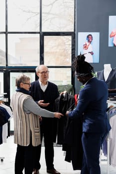 Elderly couple shopping for formal wear in showroom, asking african american employee for help with shirt. Senior man buying fashionable clothes and trendy accessories in clothing store
